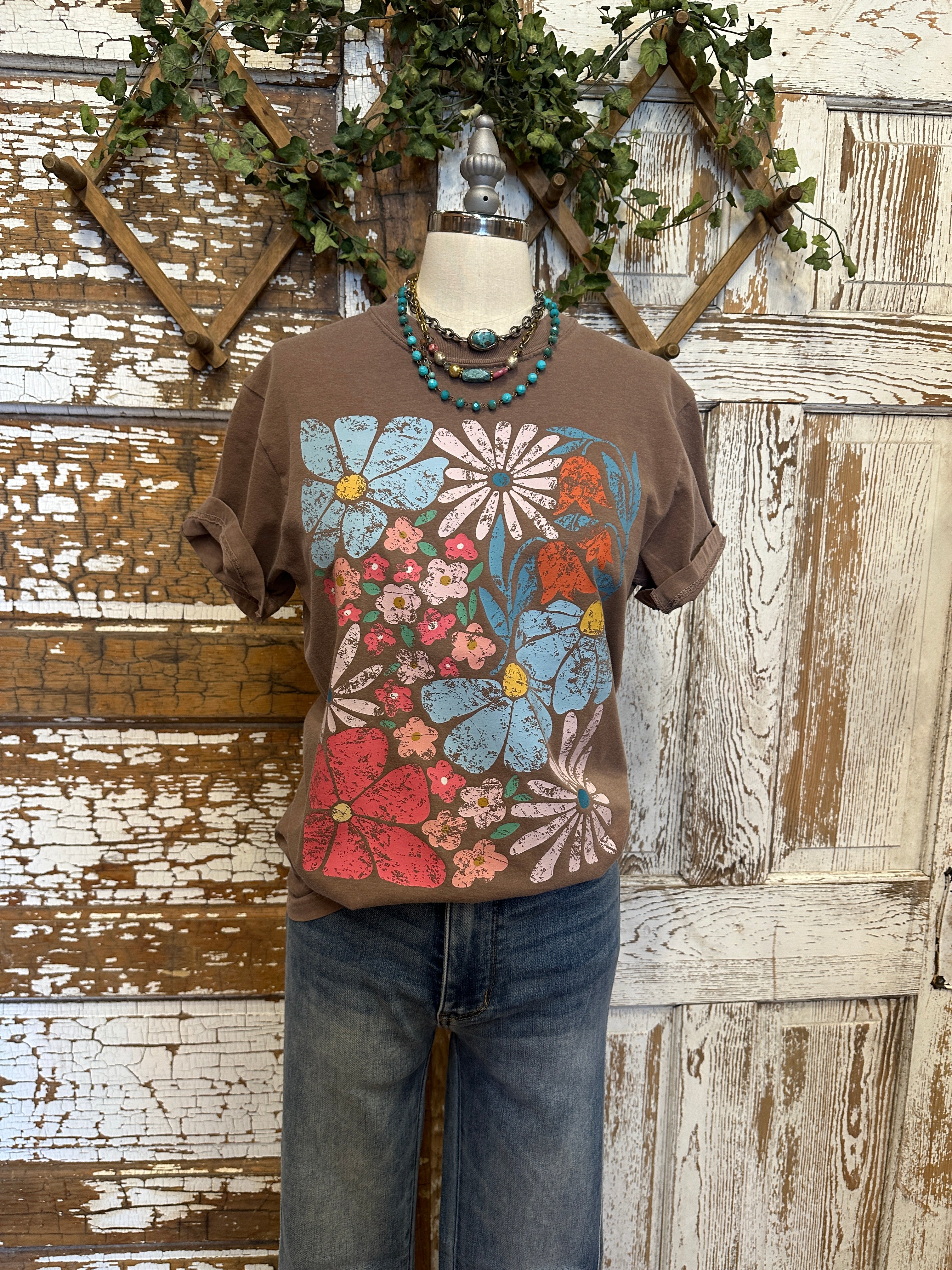 Mannequin wearing a brown graphic tee shirt with floral design and blue jeans against a rustic wooden wall.