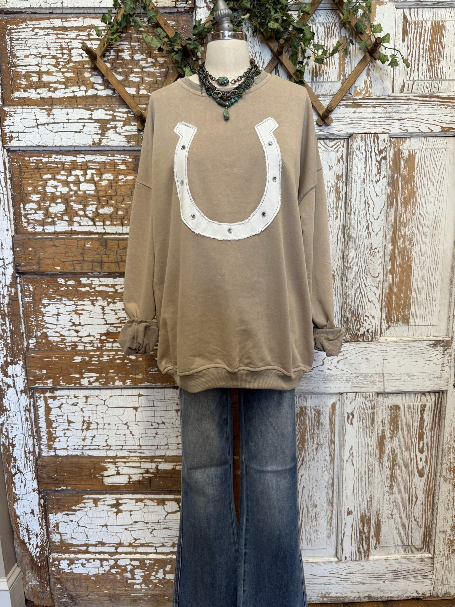 Taupe long-sleeve top with a horseshoe design on a mannequin against a rustic wooden background.