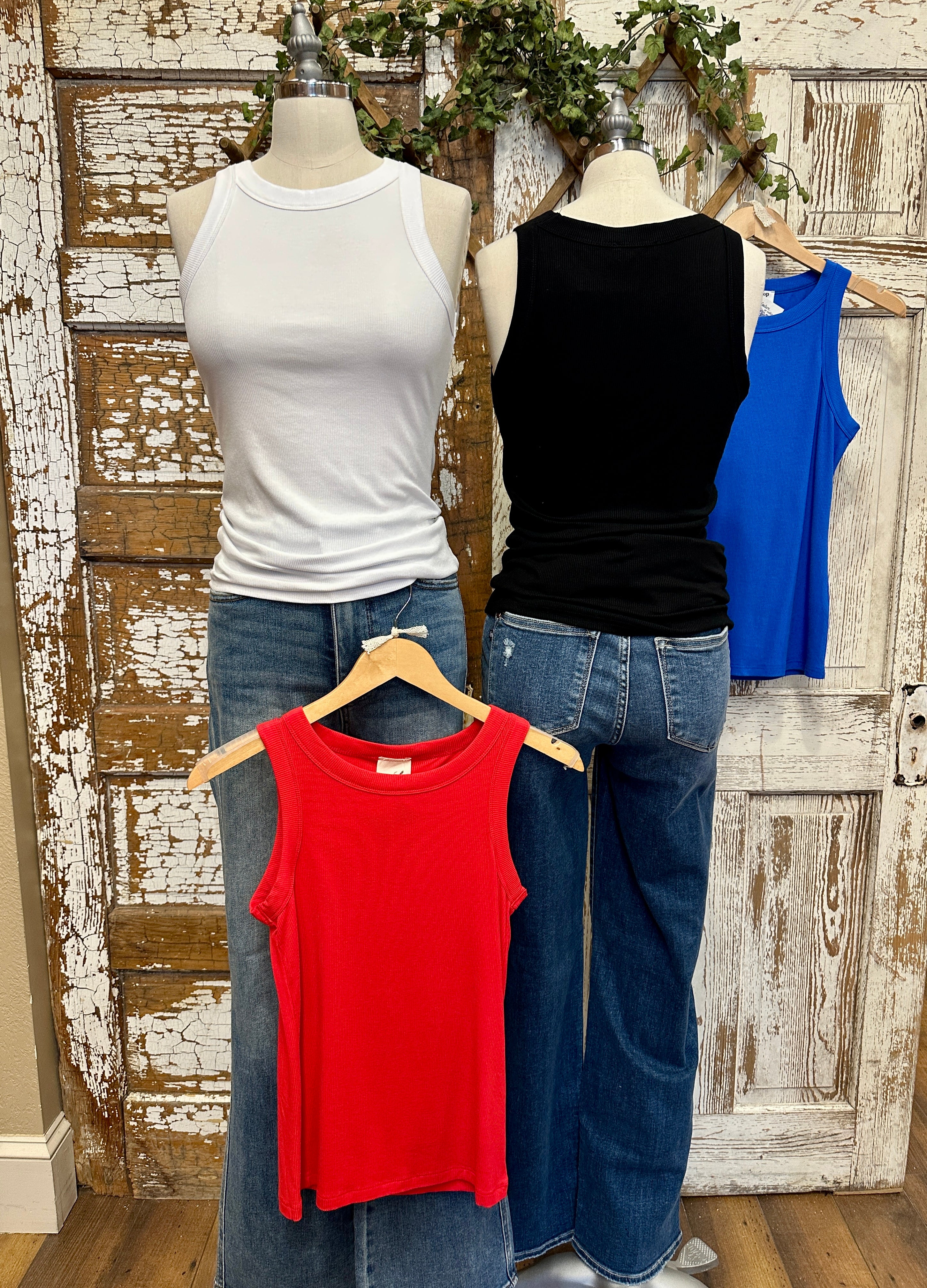 Three tank tops in blue, white, black, and red on mannequins with jeans against a rustic wooden door background.