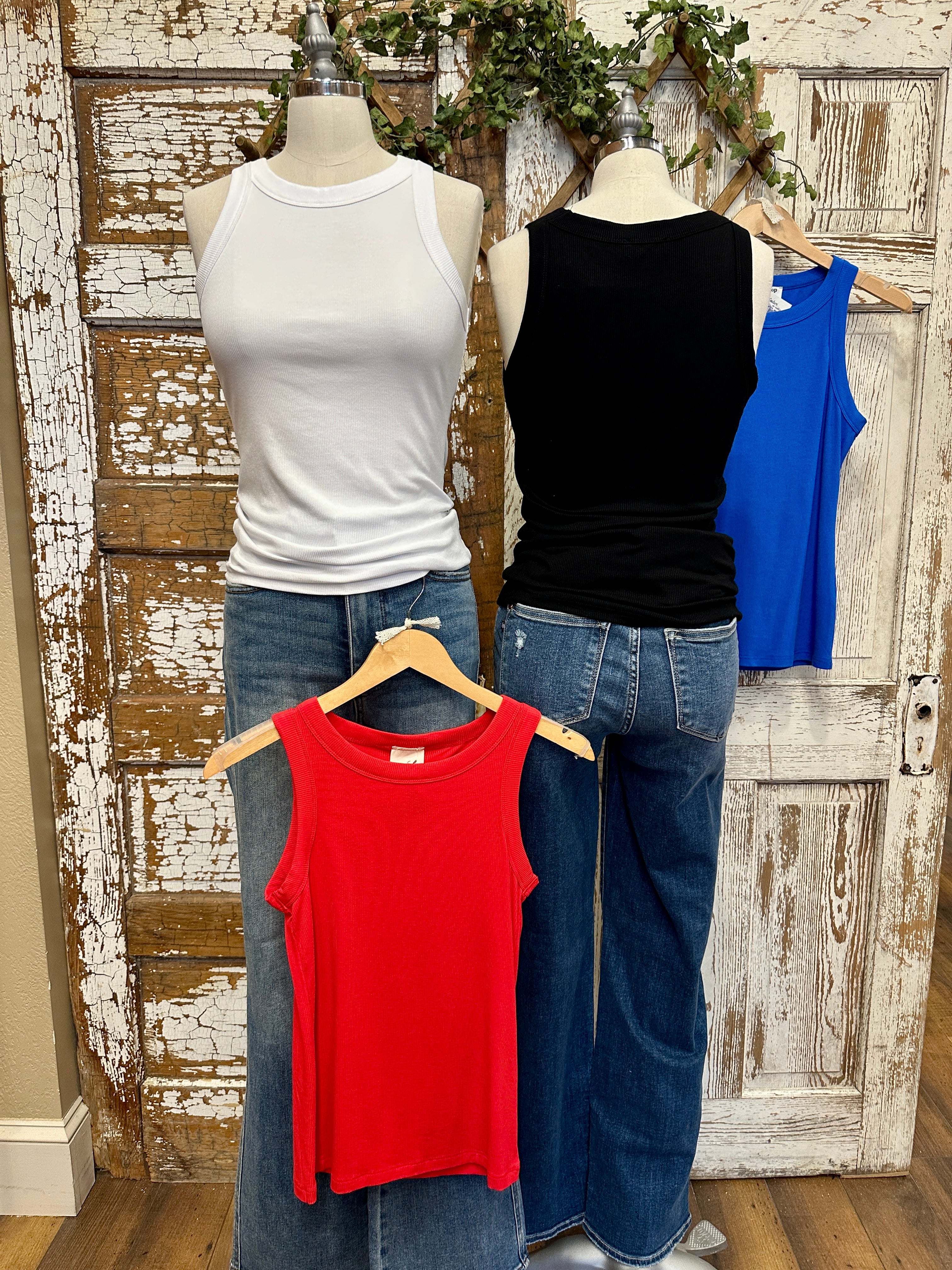 Three tank tops in blue, white, black, and red on mannequins with jeans against a rustic wooden door background.