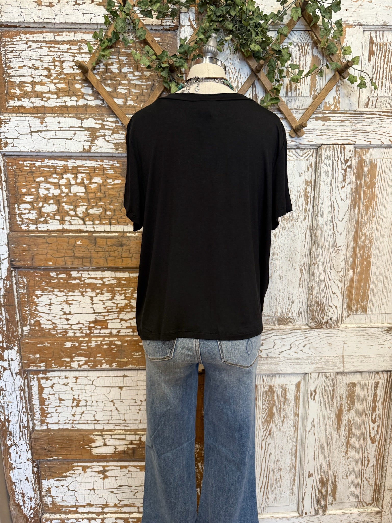 Mannequin wearing a black t-shirt and blue jeans against a rustic wooden wall with greenery.