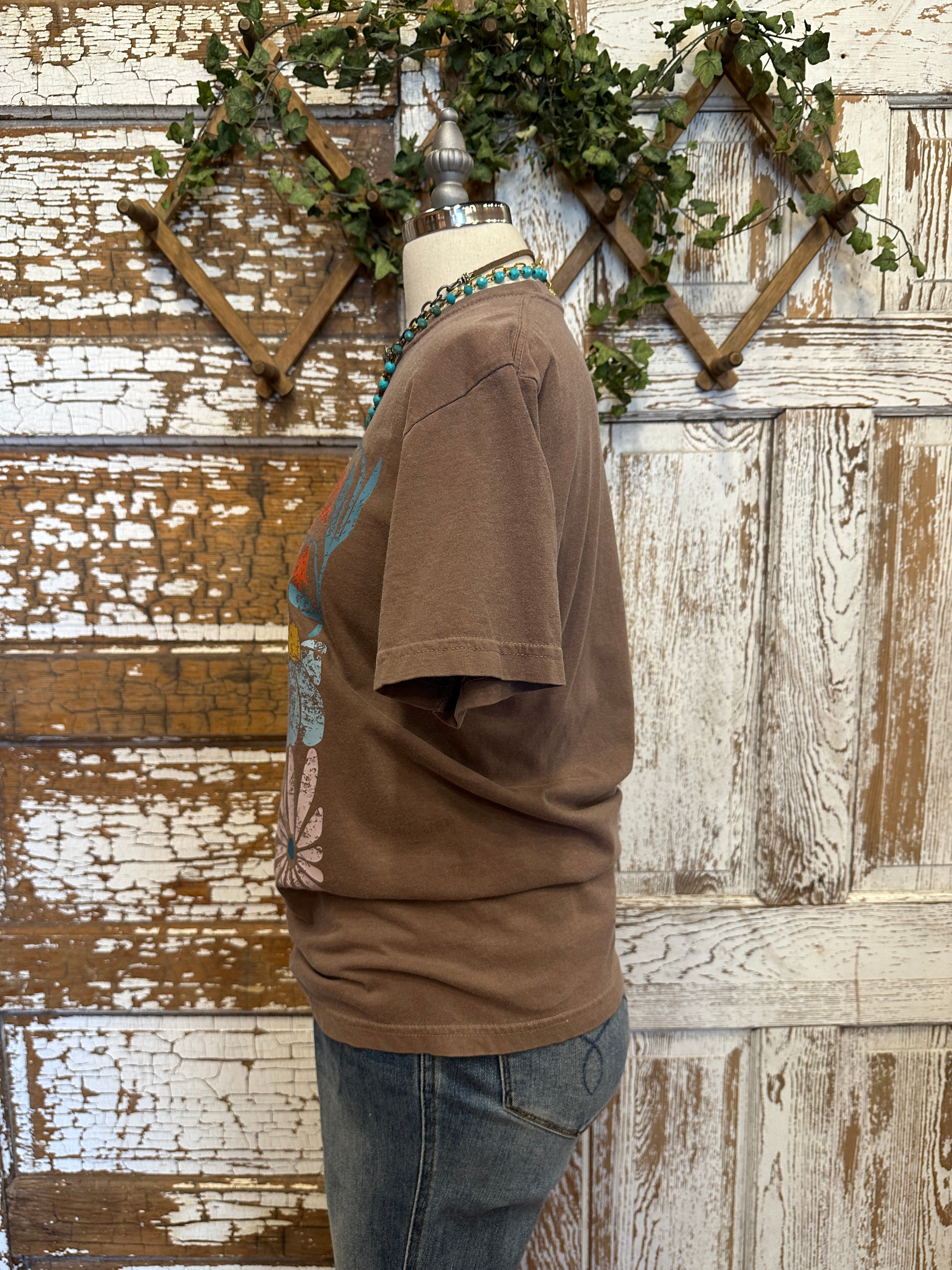 Mannequin wearing a brown graphic tee shirt with floral design and blue jeans against a rustic wooden wall.
