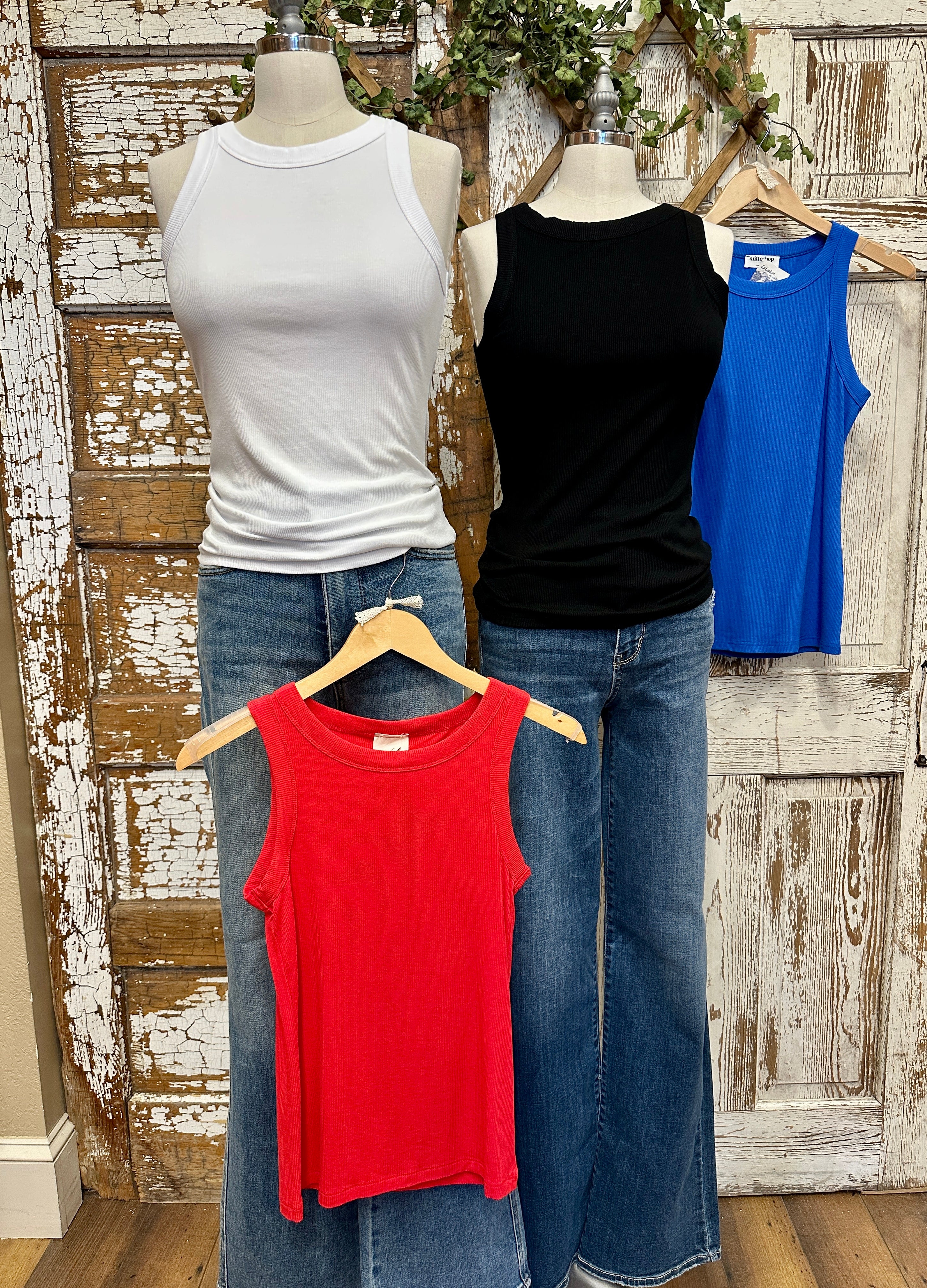 Four tank tops in white, black, blue, and red on mannequins against a rustic wooden wall.