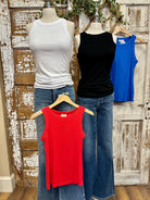 Four tank tops in white, black, blue, and red on mannequins against a rustic wooden wall.