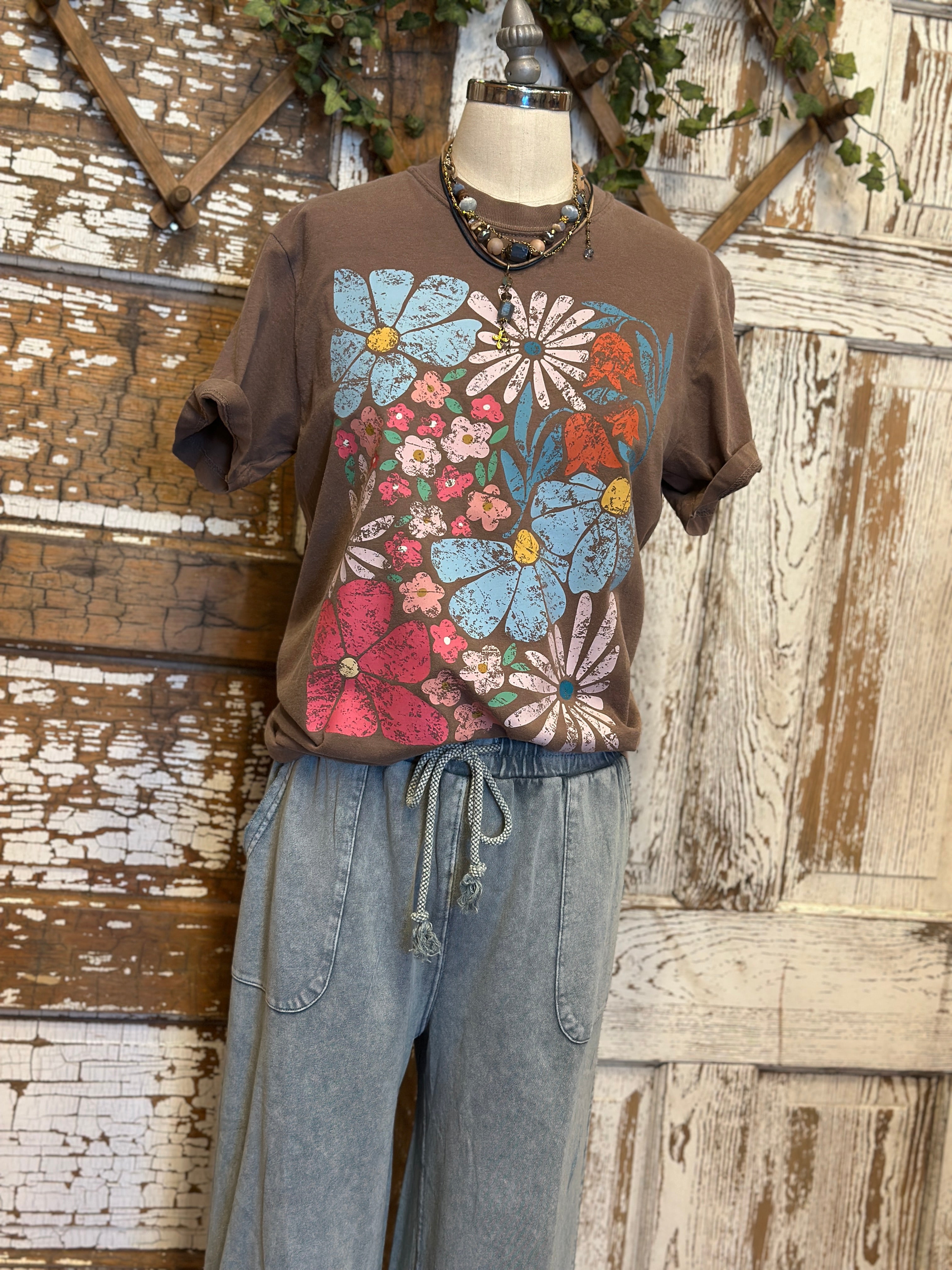 Mannequin wearing a brown graphic tee shirt with floral design and blue jeans against a rustic wooden wall.
