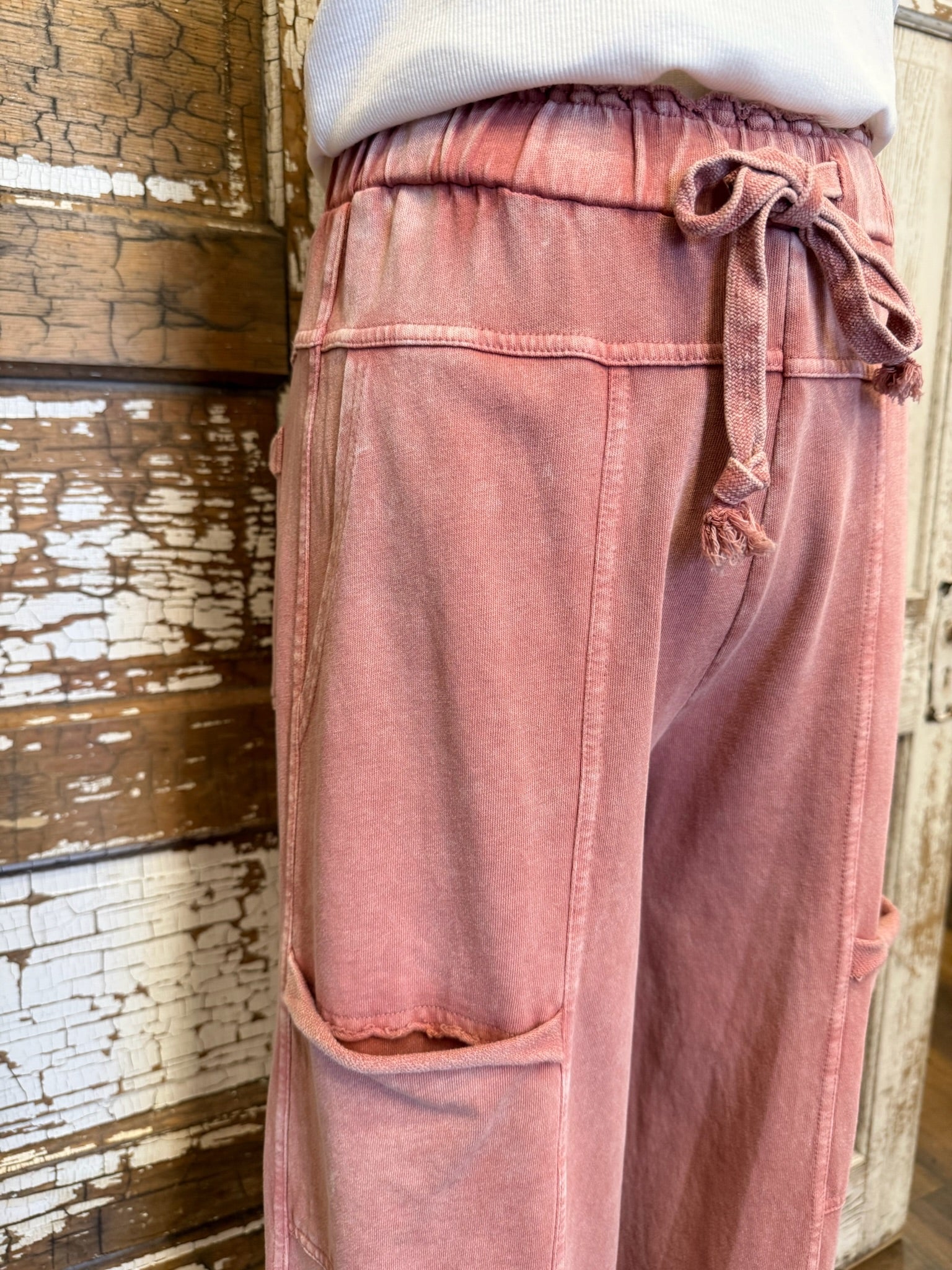 Pink pants with a tie waist worn by a person against a rustic wooden wall.