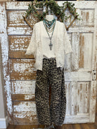 Mannequin wearing a white lace top, leopard print pants, and layered turquoise, chain and leather necklaces against a rustic wooden door background.
