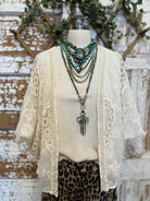 Mannequin wearing a cream lace cardigan with layered turquoise handmade necklaces against a rustic wooden background