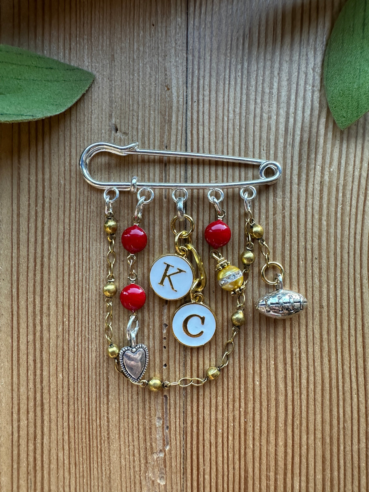 Silver-tone charm pin brooch with K and C initial charms, football, heart, red beads, golden pearl, and gold beaded chain