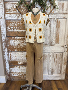 Mannequin wearing a sunflower-patterned crochet vest and beige pants against a rustic wooden door background