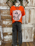 Mannequin wearing an orange graphic tee shirt with western design and gray leisurewear pants against a rustic wooden background.