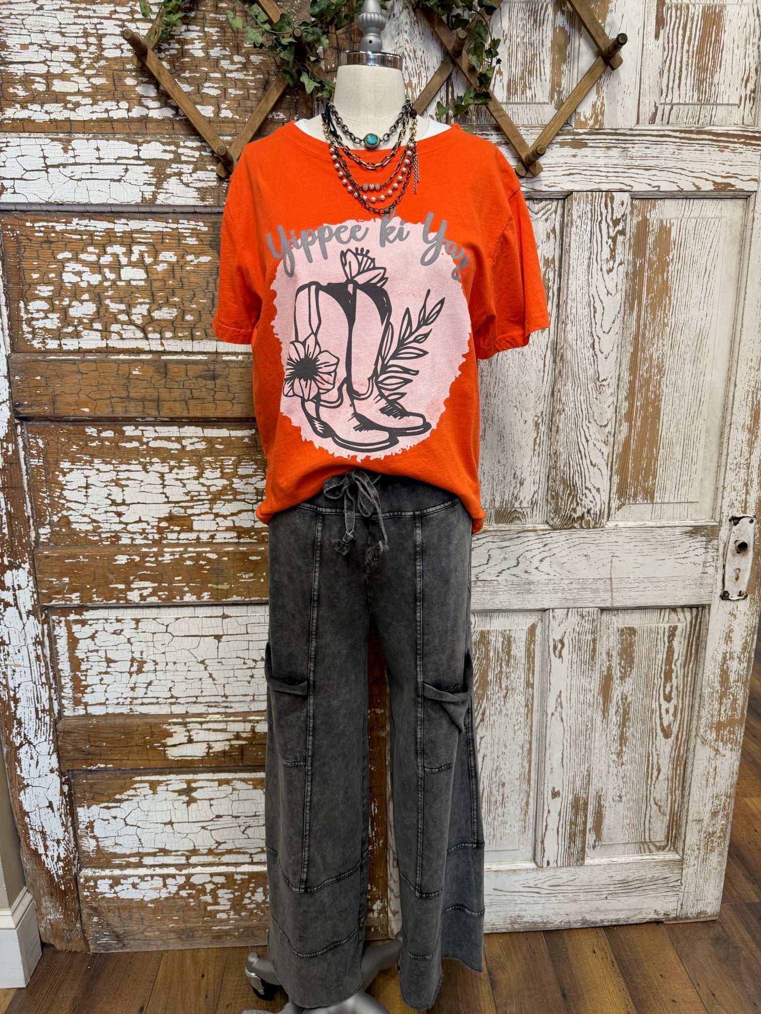 Mannequin wearing an orange graphic tee shirt with western design and gray leisurewear pants against a rustic wooden background.