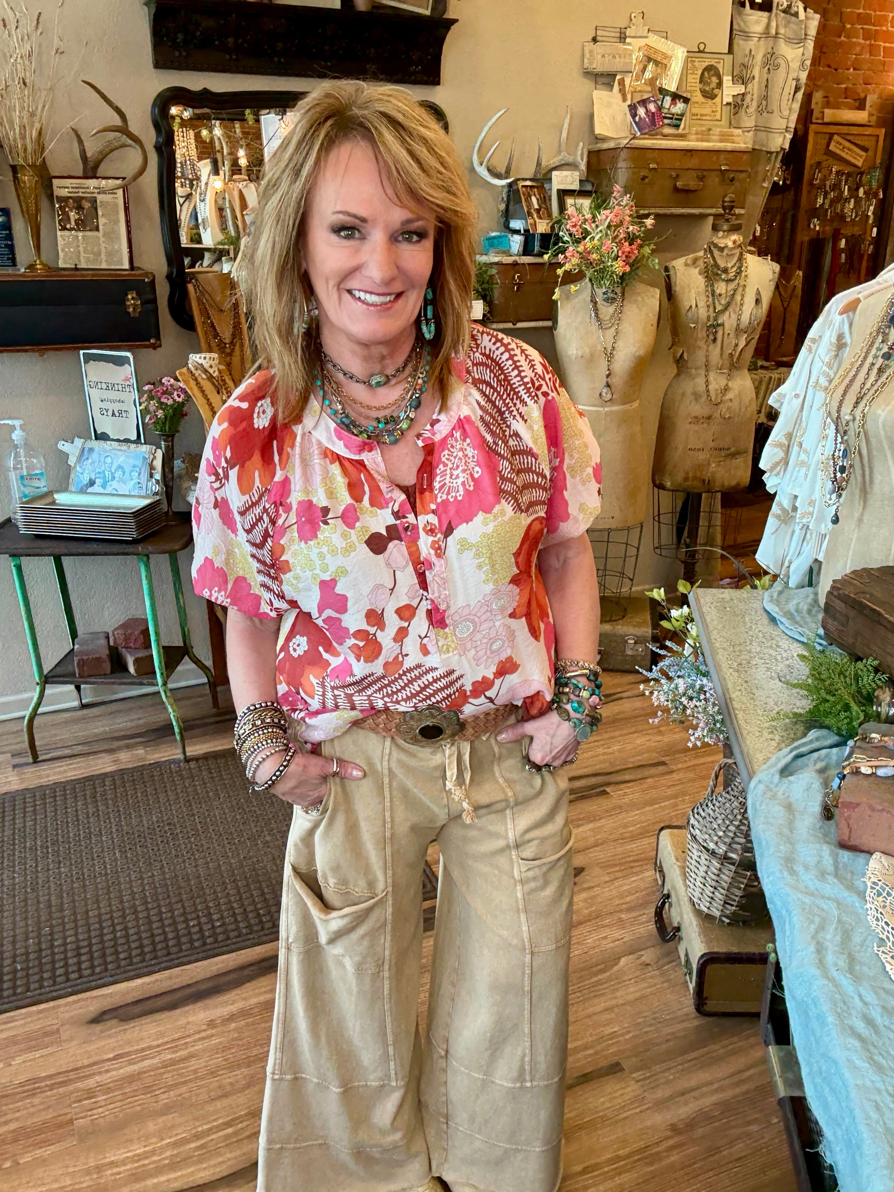 Lu wearing a floral blouse and leisurewear pants with handmade jewelry layers in her Leavenworth, KS boutique