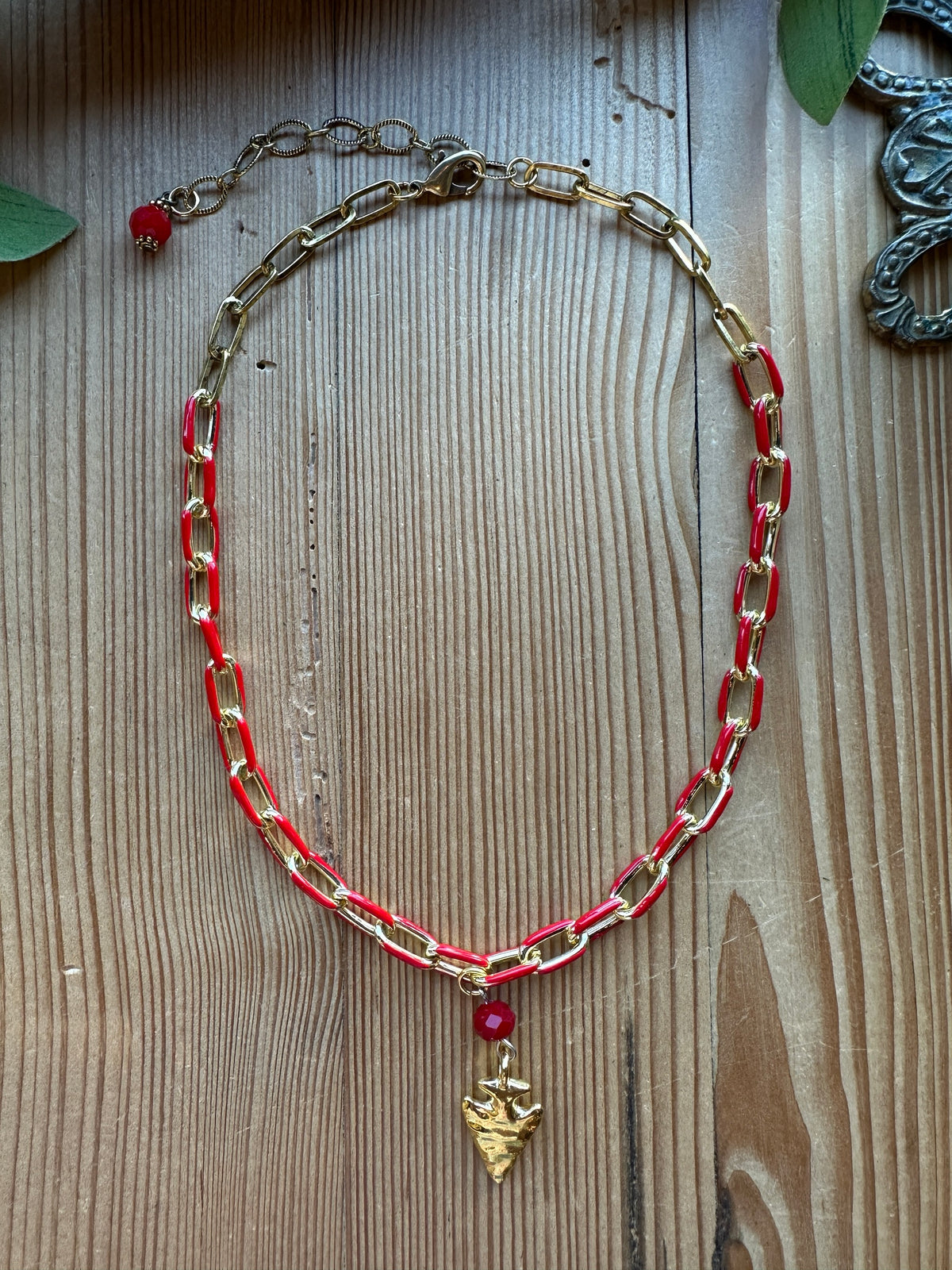 Red and gold enamel paperclip chain necklace with faceted red crystal and gold arrowhead pendant on a wood background