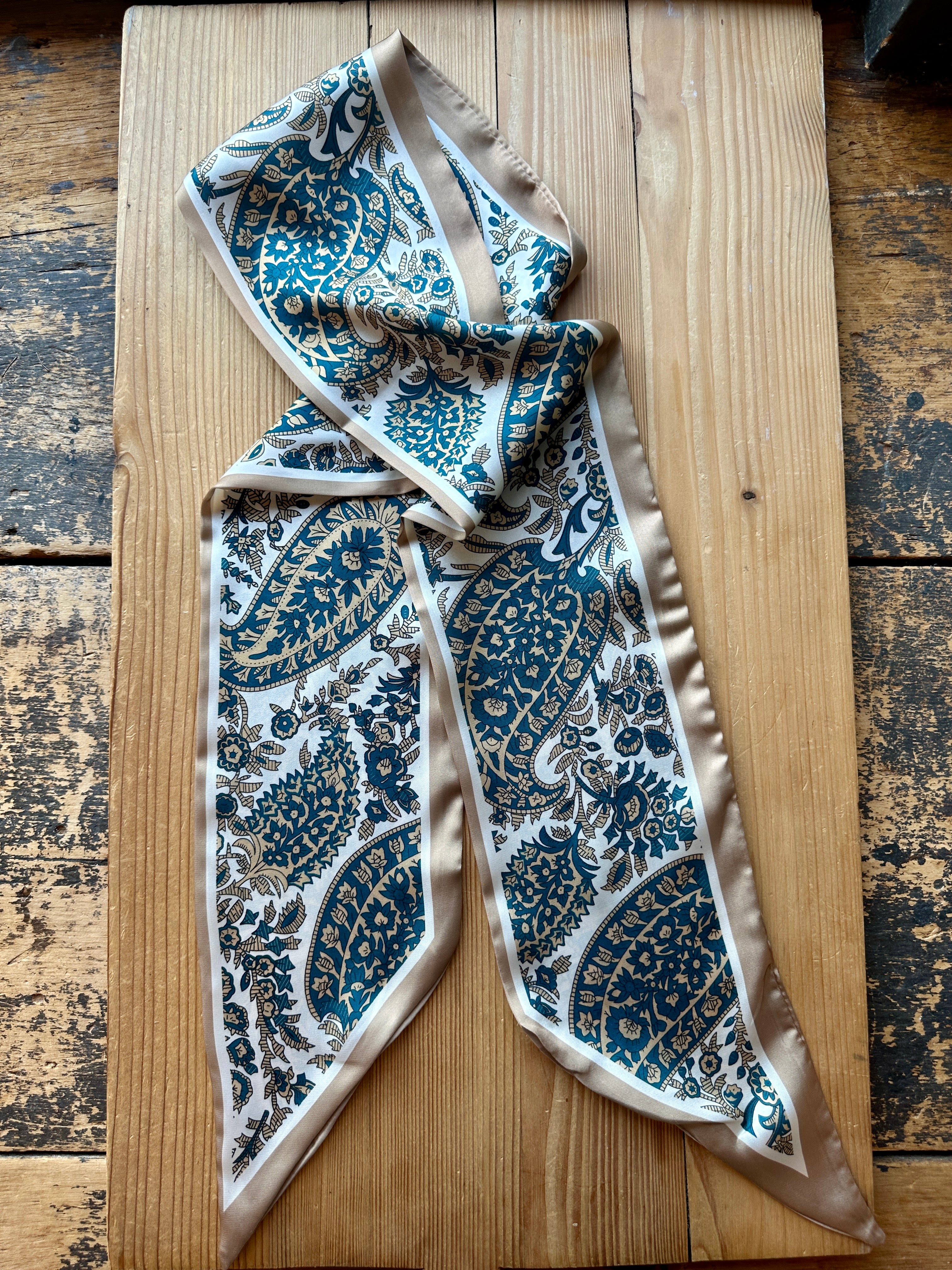 Teal paisley print silky twilly scarf laid flat on a wood surface
